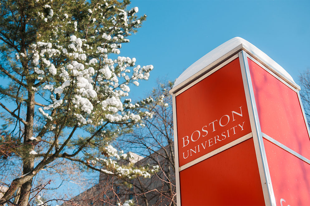 Boston University sign with snow on top in front of a snowcovered tree