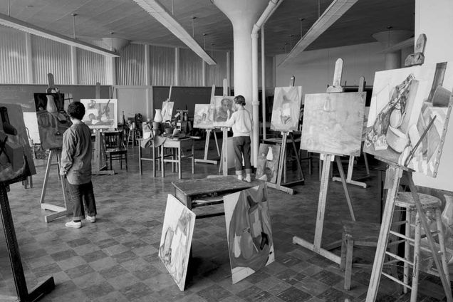 black and white photo: painting studio with many canvases on easels in various states of completion, and two currently being worked on by students.
