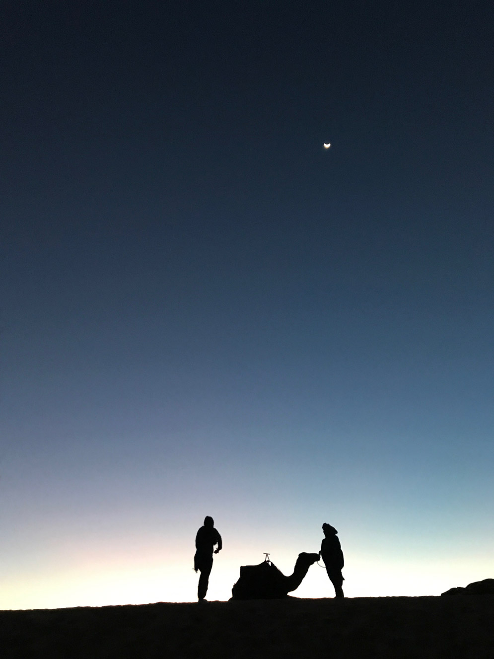 silhouette of two people and a camel on a sand dune with the sun about to rise, the moon is in a heart shape