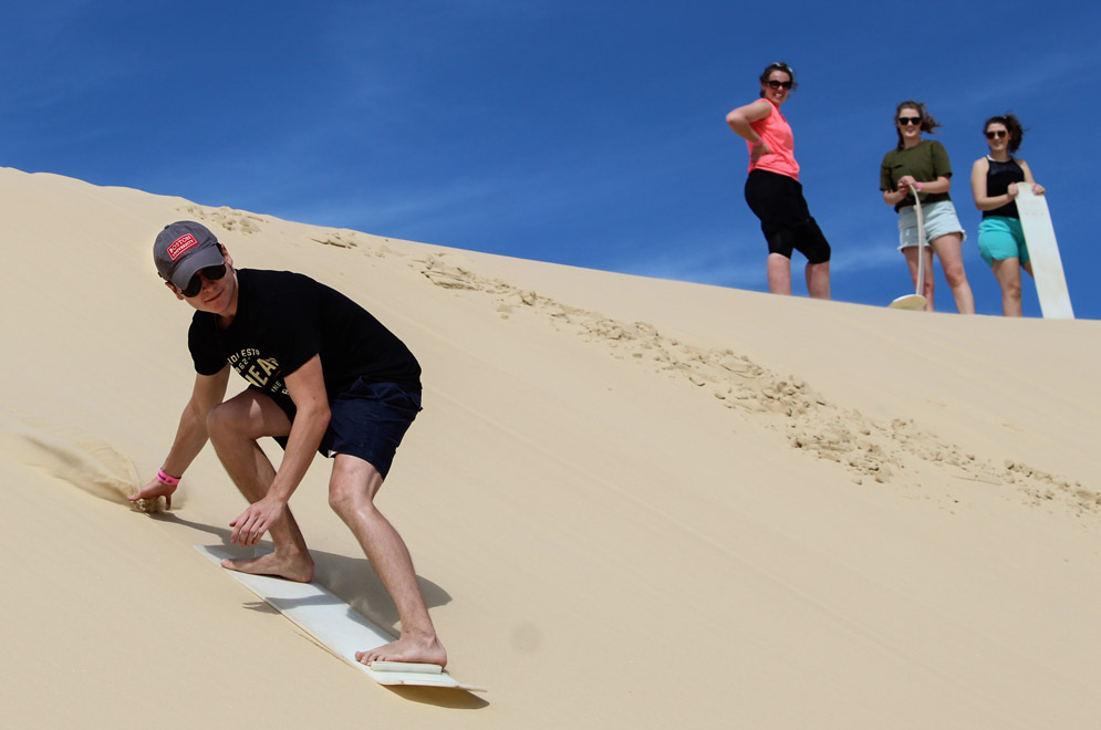 A student wearing a Boston University hat boards down a sand dune with three others watching from the top