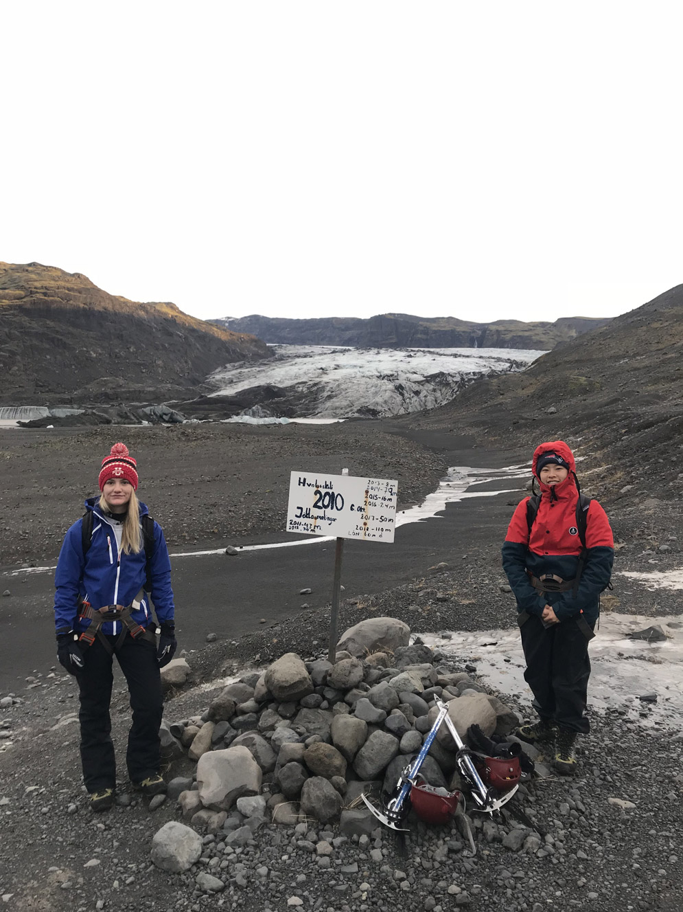 two students, wearing warm clothing and Boston hats, stand next to a pile of rocks with hiking gear and a sign that has dates and distances on it