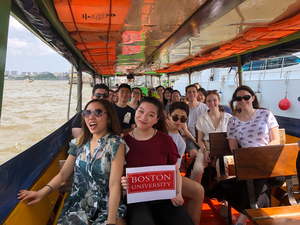 people sitting on a boat with a city in the distance; one person is holding a Boston University sign