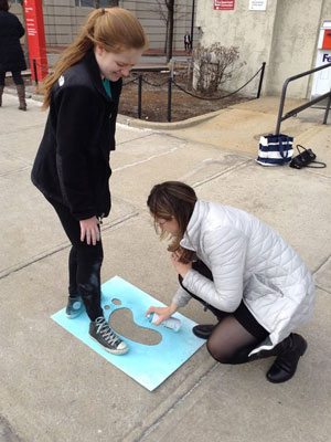 Russell standing on a stencil while another student spray paints the sidewalk