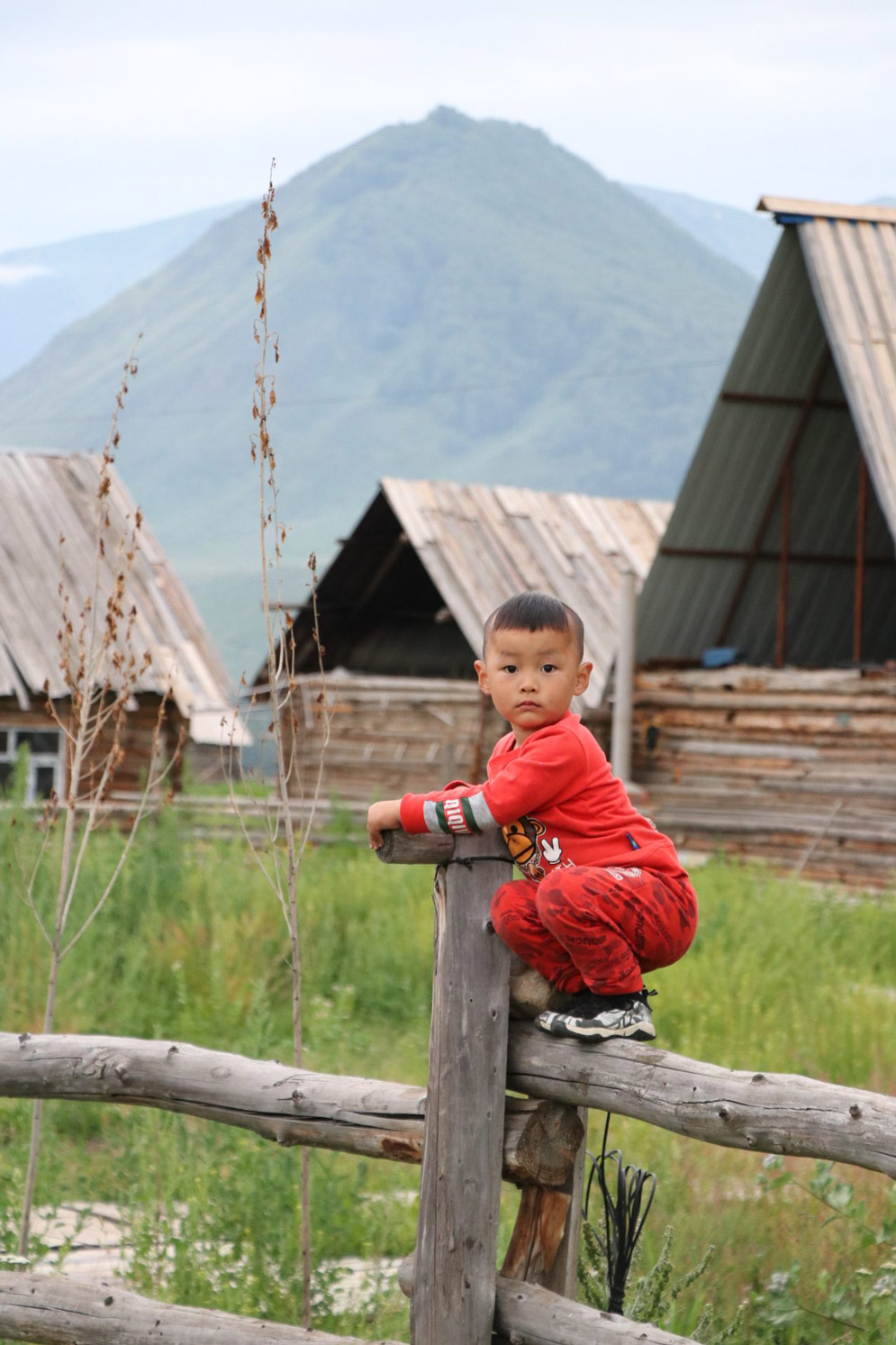 a kid in a red outfit sits on a log fence, with log houses in the background