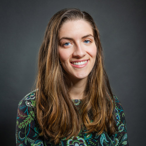 Photo of Jessica Colarossi. A white woman with long, straight brown hair and wearing a black and green paisley blouse smiles and poses in front of a dark grey background.