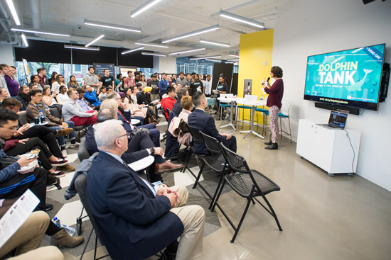 Ziba Cranmer stands in front of an audience next to a screen that reads "DOLPHIN TANK BUILD LAB *Demo Day" along with listing some sponsors and other details and showing a web link to vote