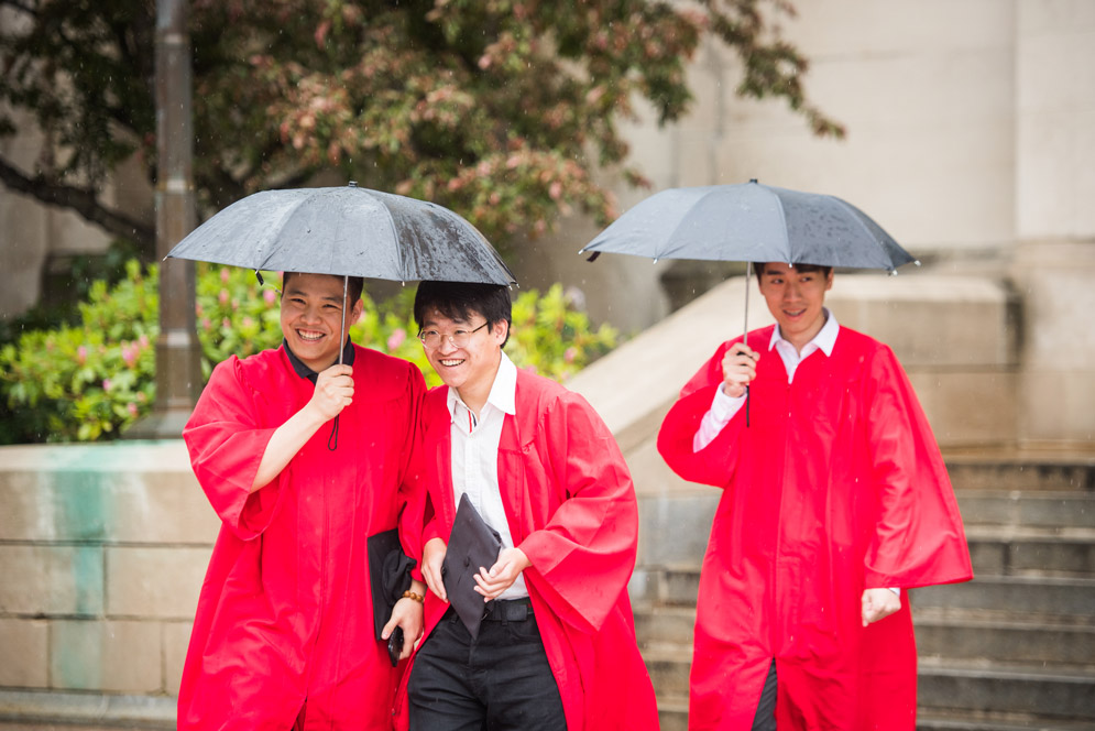 Three boys walk under umbrellas in their commencement gowns