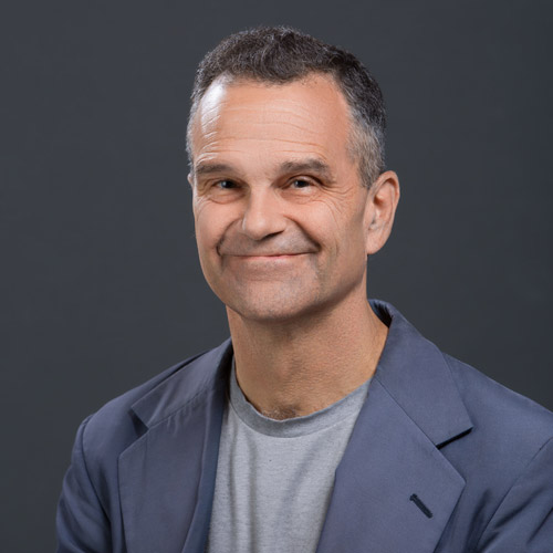 Photo: Headshot of Rich Barlow, an older white man with dark grey hair and wearing a grey shirt and grey-blue blazer, smiles and poses in front of a dark grey backdrop.