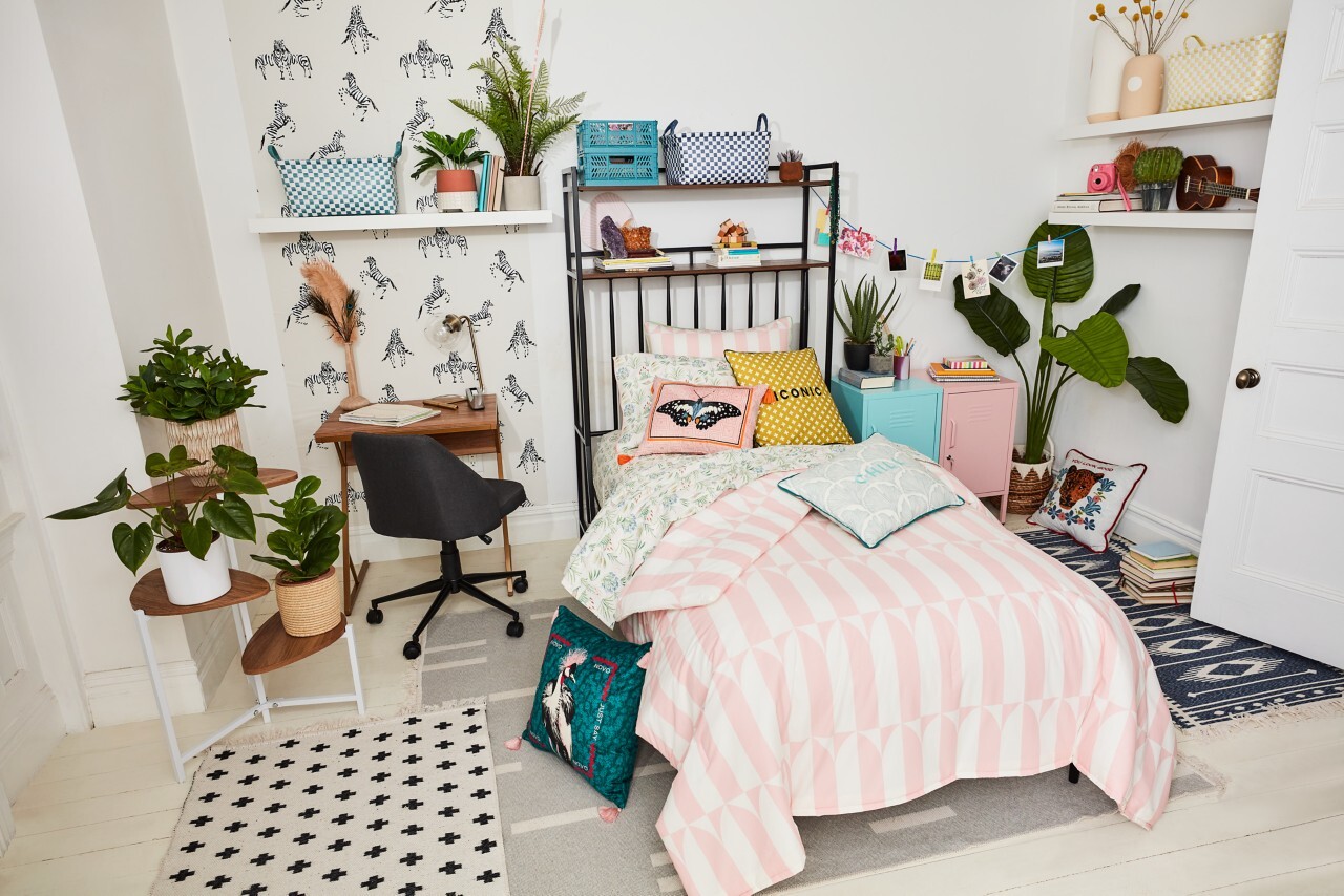 Photo of a dorm room decorated in pastel colors and plants. A small desk is shown next to a twin size bed with plants and knick knacks used to decorate rom.