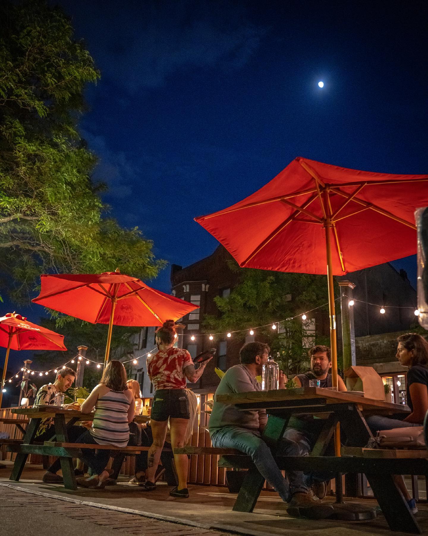 Photo of Blossom Bar’s patio filed with people at night with the moon seen in the dark sky above them. The tables have red umbrellas and a server brings food to one of the tables.