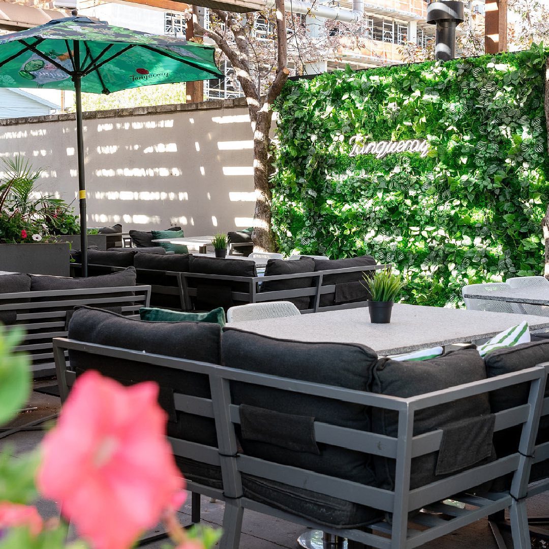 Photo of the light and dark gray tables and chairs on the Earls patio; one of the wall is decorated with fake greenery and features a neon Tanqueray sign.