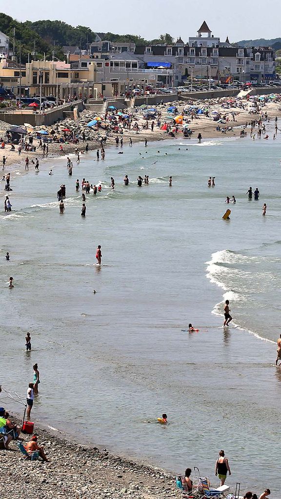 Wide photo of visitors crowding onto Nantasket Beach in Hull, MA. People are seen sitting on the small, partially rocky shore at left, while others are in the shallow water at right. Houses and buildings lining the street along the beach are seen in the background.