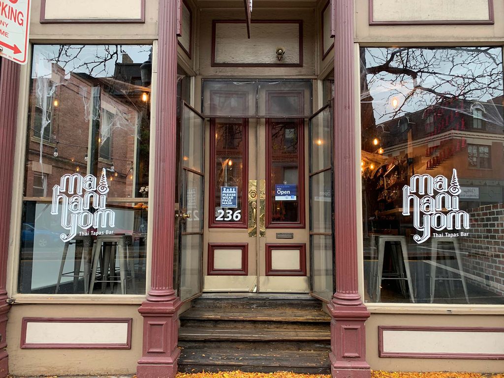 Photo of the exterior of Mahaniyom. The white logo is seen in both windows on either side of the door; tables and stools are seen inside.