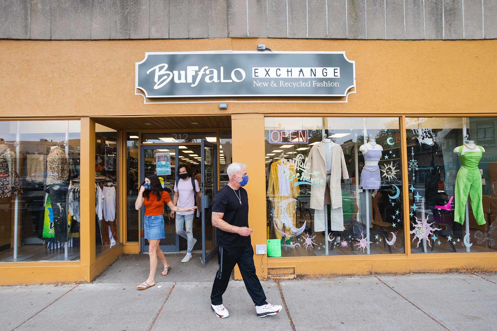 Photo of the exterior of Buffalo Exchange (312 Harvard St.). Three people with face masks walk out of the orangey brown store front that has a black and white sign.