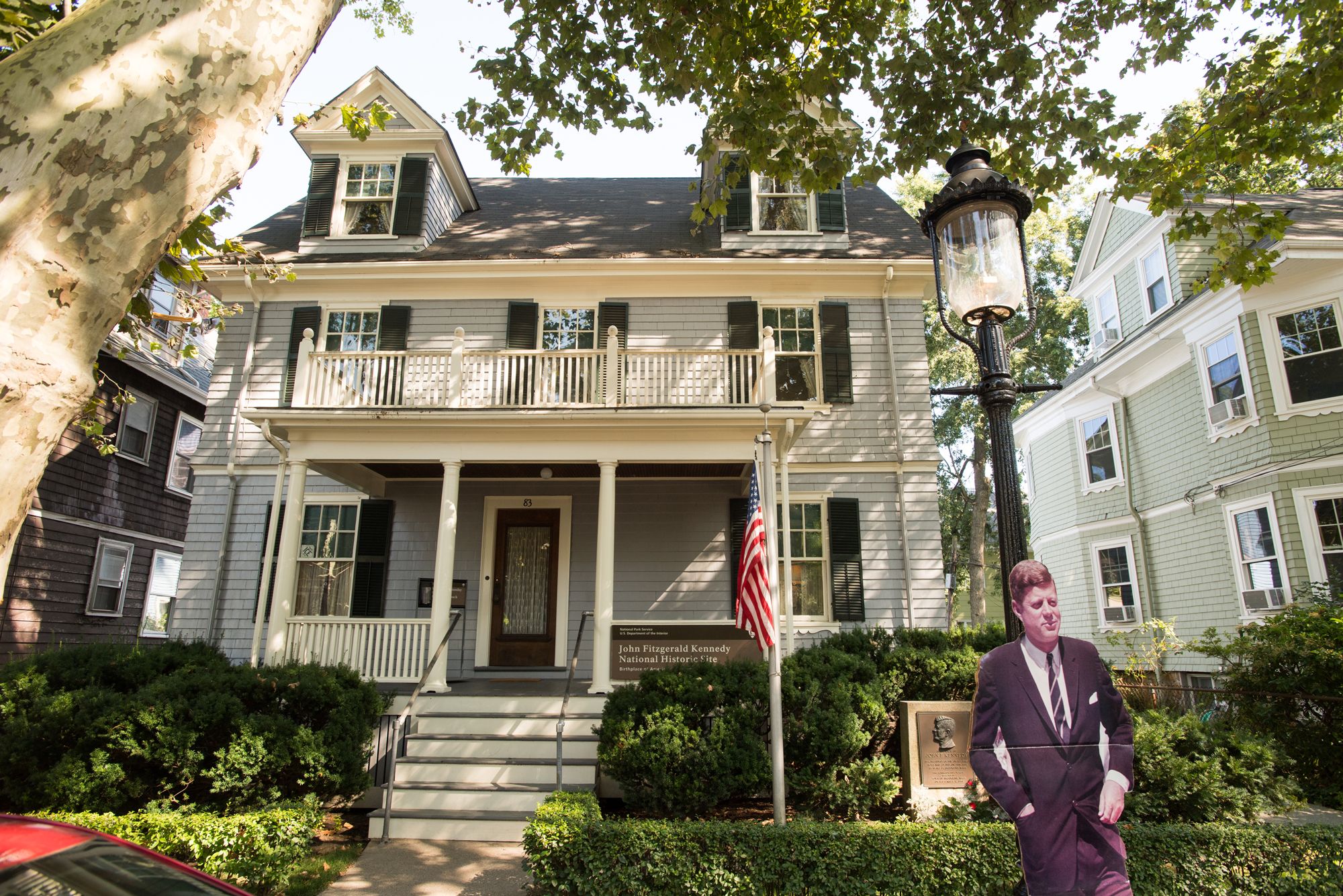 Photo of a the JFK house. A grey house is shown n a suburban street. A cutout of JFK is placed in front of the house.