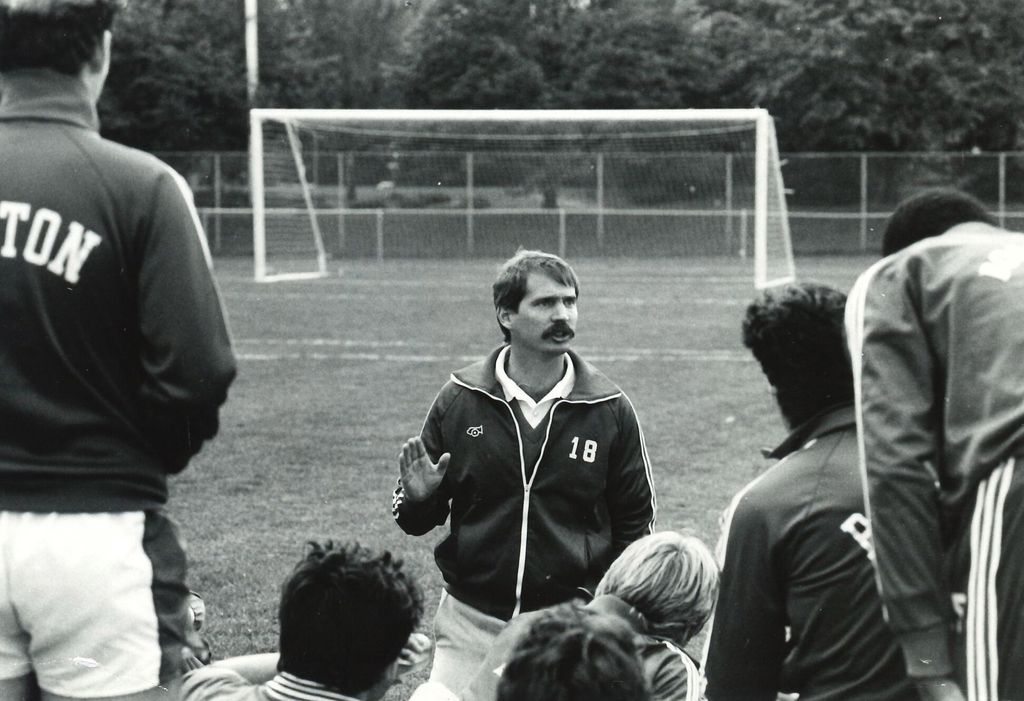 Neil Roberts talks to his team while kneeling on the field. A soccer goal frames him
