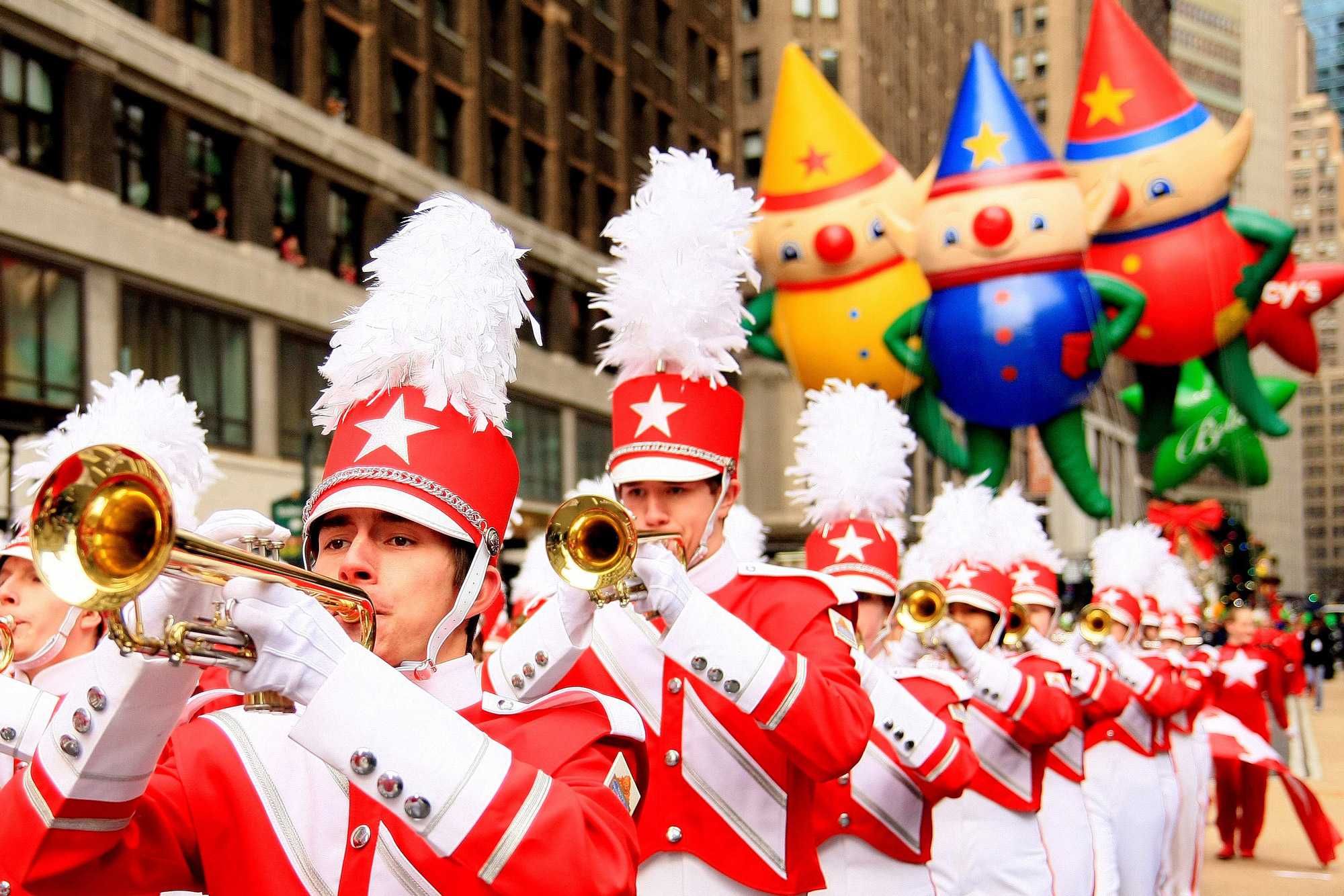 A group of trumpet players marches during the Macy's day parade with balloons floating in the distance