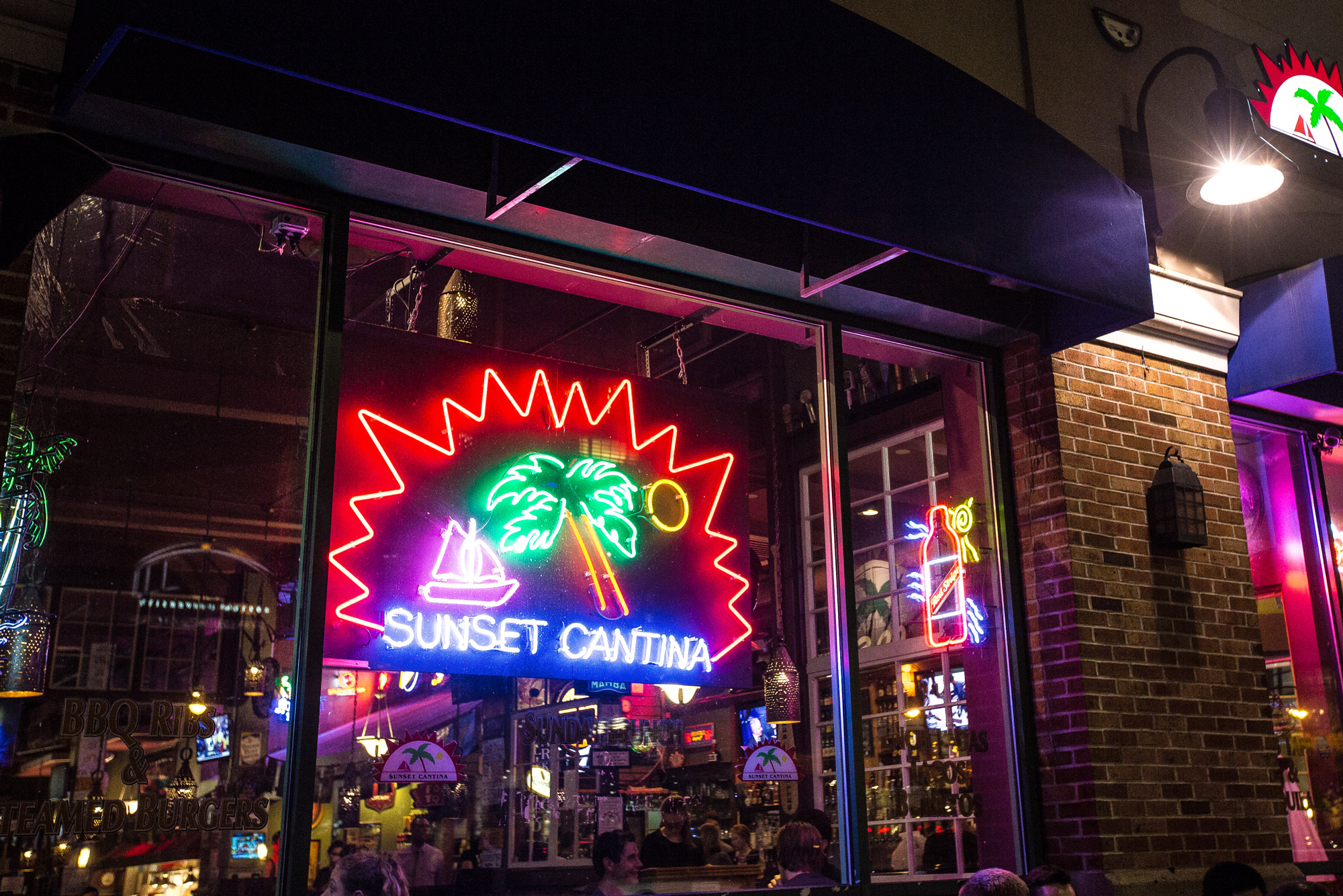 A colorful neon sign hanging in Sunset Cantina's window spells out "Sunset Cantina" with a sailboat and palm tree during night-time