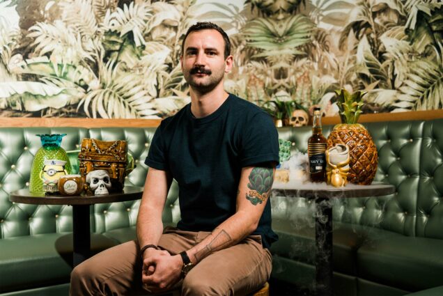 Portrait of Ryan Lotz, Beverage DIrector at Shore Leave Boston tiki bar, sitting in a green banquette surrounded by tropical wallpaper and tiki culture decoration. Photo by Chris McIntosh
