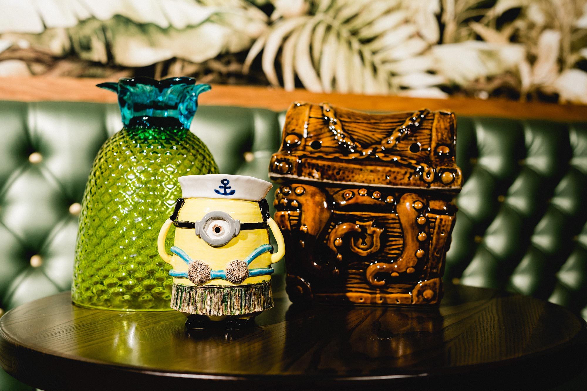 Tiki decoration including a glass pineapple shaped bowl, Despicable Me Minion dressed in a hula skirt and sailor cap, and ceramic treasure chest with octopus tentacles sticking out of it on a table at Shore Leave tiki bar in Boston.