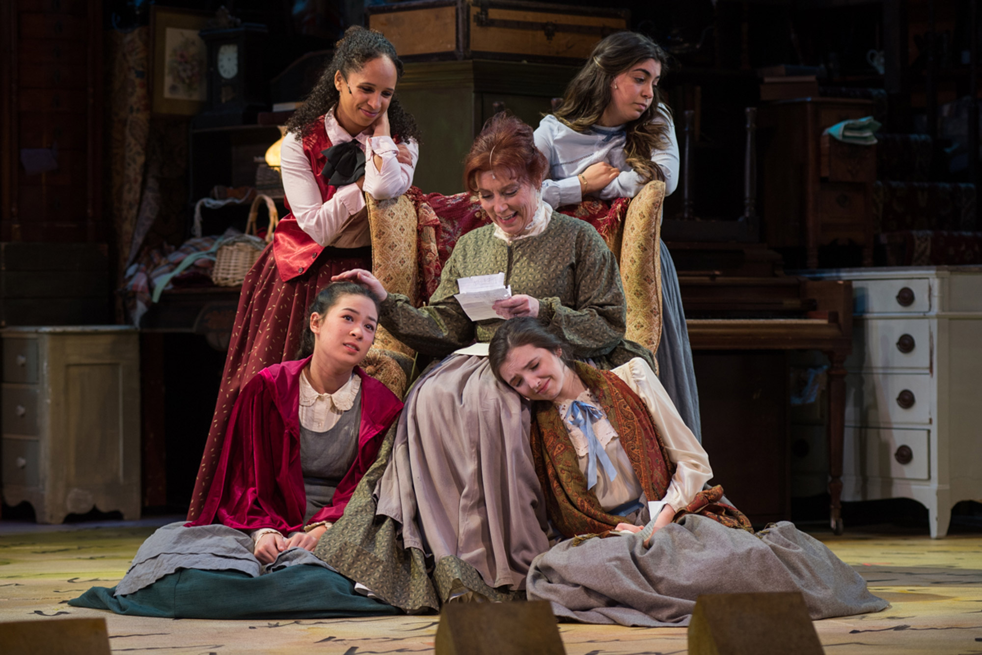 A photo of the "Little Women: The Broadway Musical" production with the characters Jo, Amy, Beth, Meg and Marmee reading a letter.