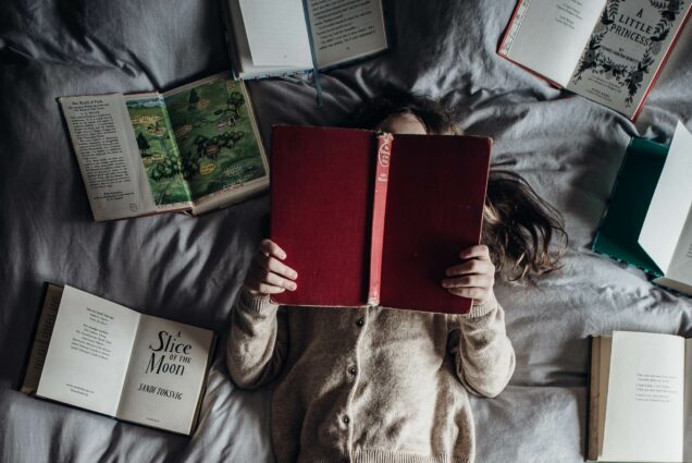 A photo of a woman on a bed reading a book surrounded by other open books.