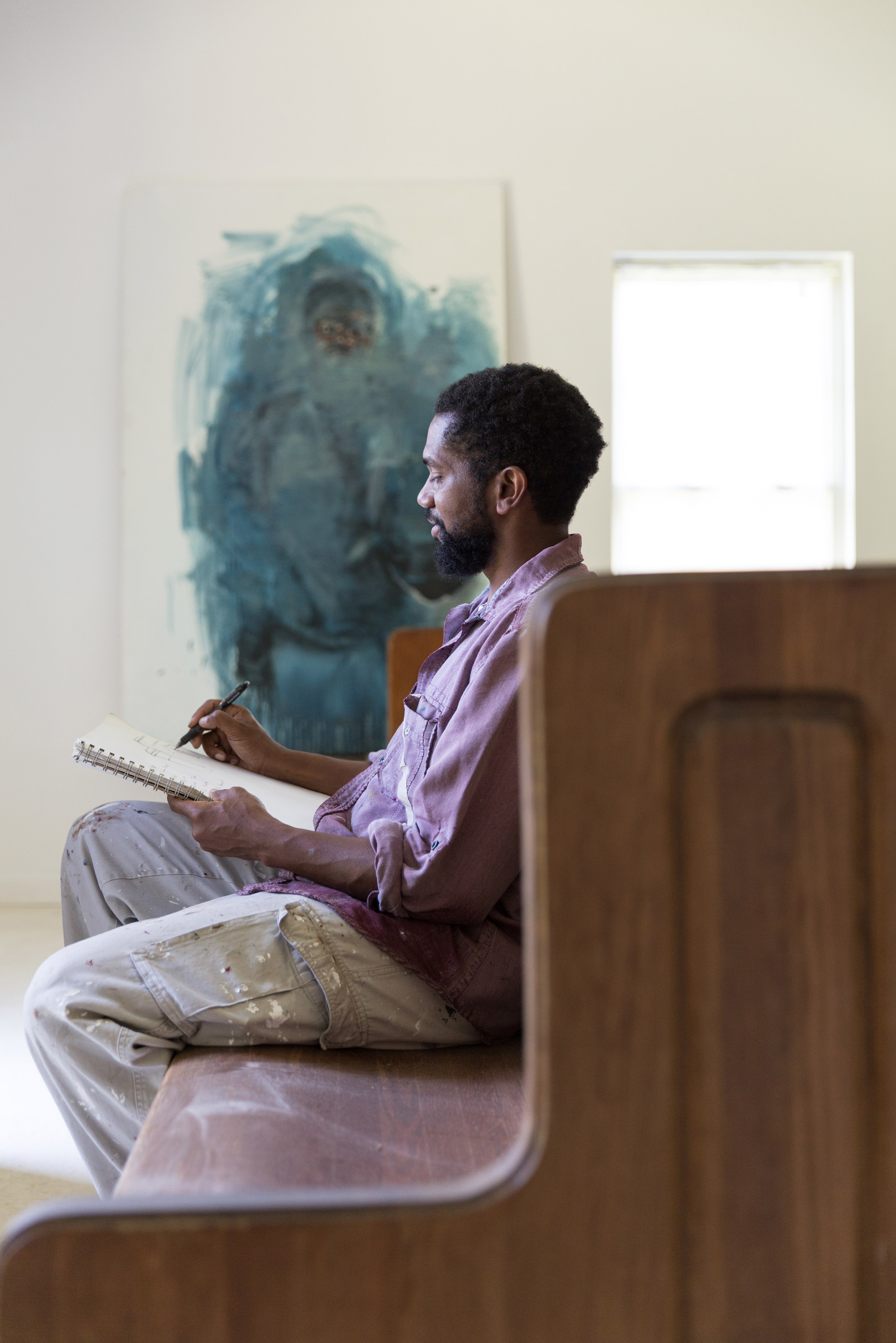 A portrait of painter Sedrick Huckaby sitting on a bench and sketching.