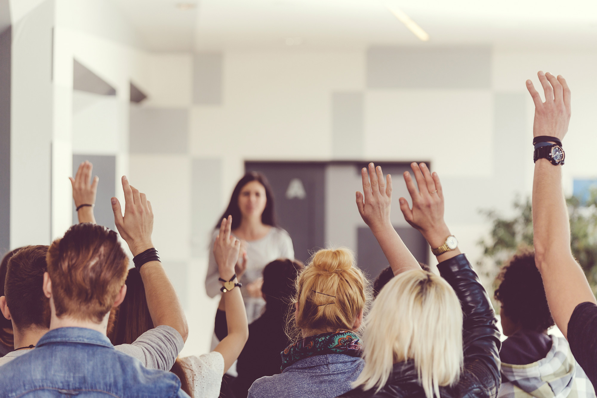 Classroom of students with hands raised, with instructor out of focus in the background.