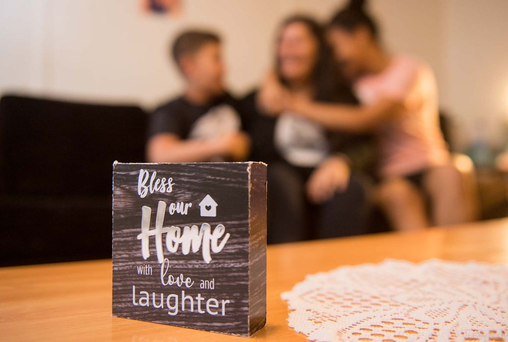 Maria's children hug her sitting on a couch behind a sign that says 'Bless our home with love and laughter.'