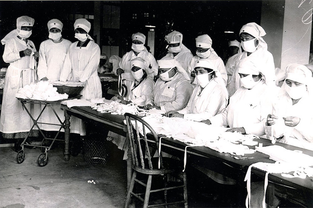 Black and white image of a group of nurses in white with facemasks. These Boston nurses are prepping masks for soldiers at Camp Devens during 1918 flu pandemic.