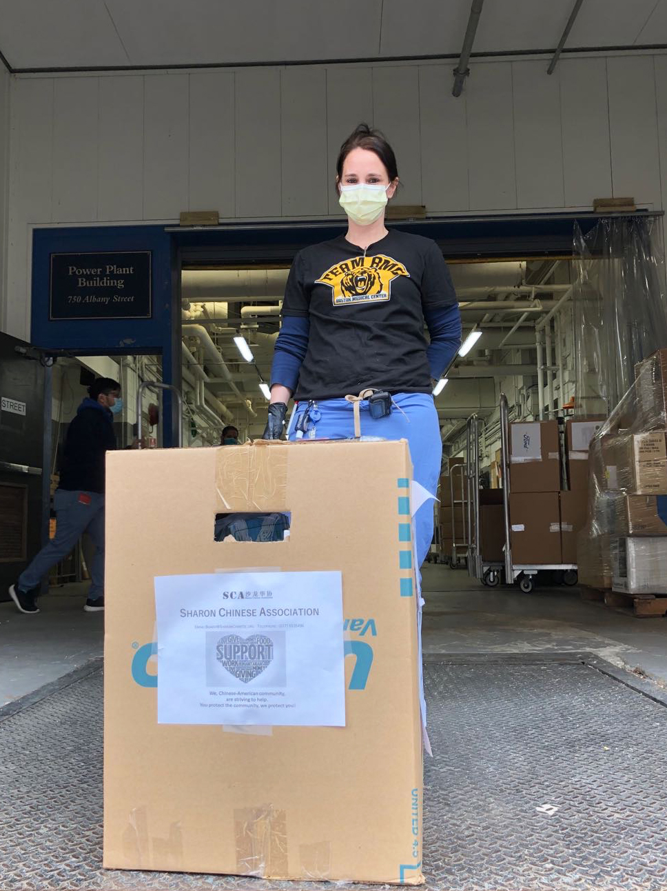 A woman with a mask and Bruins t-shirt on standing on a loading dock with a cardboard box in front of her that says “SCA - Sharon Chinese Association.”