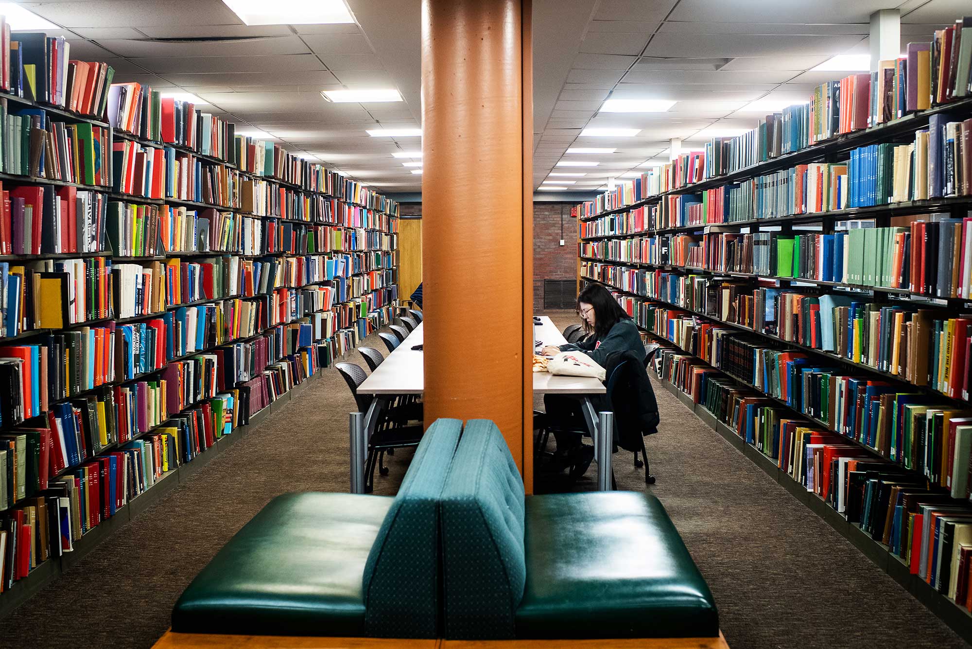 Image of a studtent studying at the Mugar Library during the campus quarantine on March 2020. There is a green couch in the foreground, and colorful bookshelves on either side of the student.