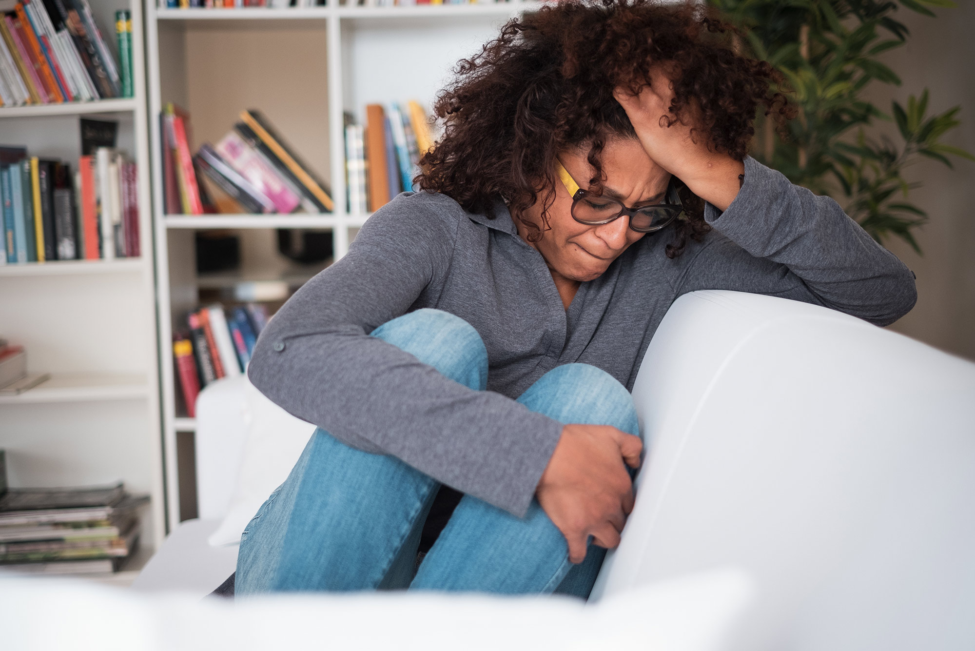 Image of a women sitting on her couch; she has her head in her hand and looks distressed.