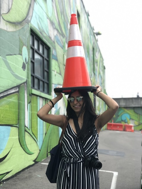 Photo of Gayatri Bajaj (Sargent’20) with a traffic cone held over her head and a mural in the background.