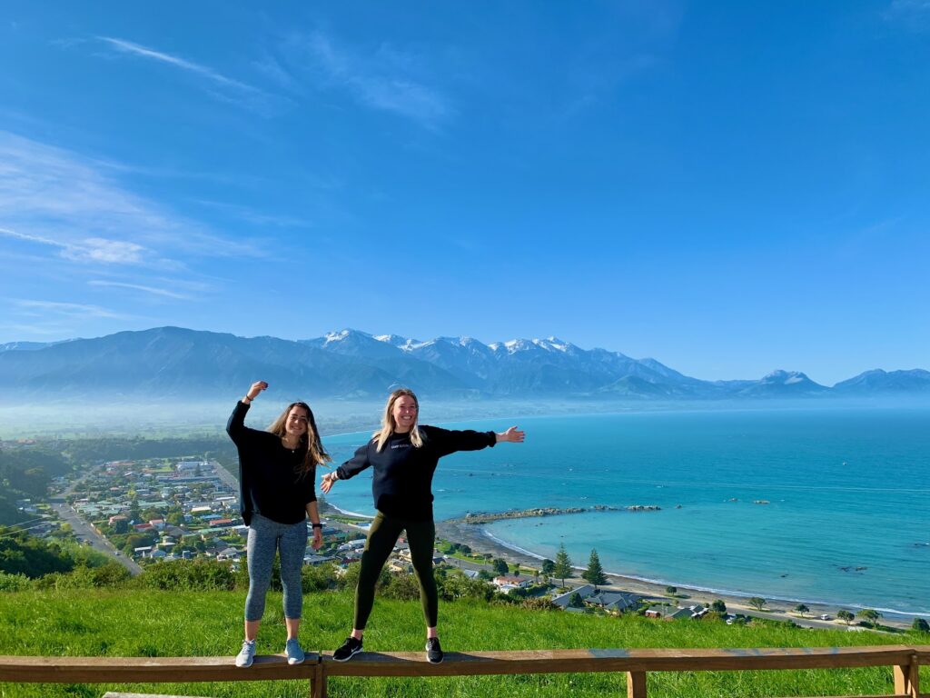Photo of Lexi Ewanich (Questrom’20) with a friend, standing on top of a rail, with a blue sky, ocean and mountains in the background.