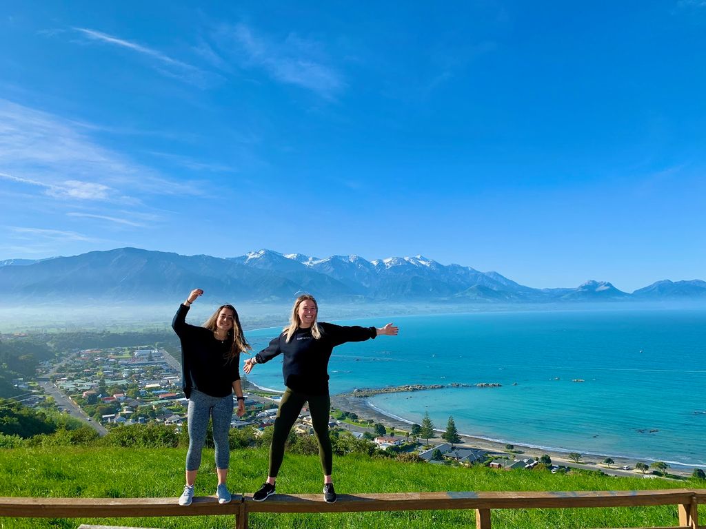 Photo of Lexi Ewanich (Questrom’20) with a friend, standing on top of a rail, with a blue sky, ocean and mountains in the background.