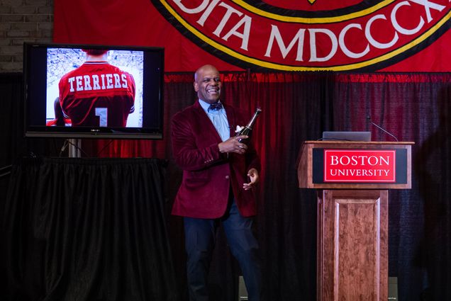 Dean Kenneth Elmore stands on a stage in the George Sherman Union next to a podium with a red Boston University sign on it. He wears a red blazer and holds a champagne bottle. Behind him, a screen shows an image of the back of a Terriers jersey.