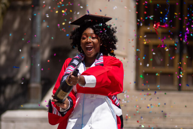 Ann-Lyssa Asare (Pardee’20) pops a champagne bottle full of confetti for a photo shoot in Marsh Plaza May 13, 2020. She wears a graduation cap and a red robe.