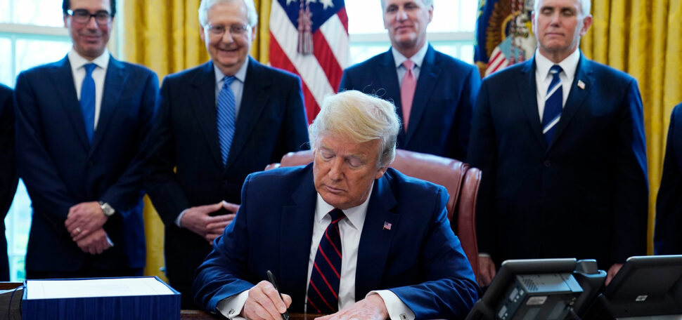 Image of President Donald Trump as he signs the coronavirus stimulus relief package, at the White House, Friday, March 27, 2020, in Washington DC. In the background, from the left, stands Treasury Secretary Steven Mnuchin, Senate Majority Leader Mitch McConnell of Ky., House Minority Kevin McCarthy of Calif., and Vice President Mike Pence, look on. All the men are wearing suits, some are smiling.