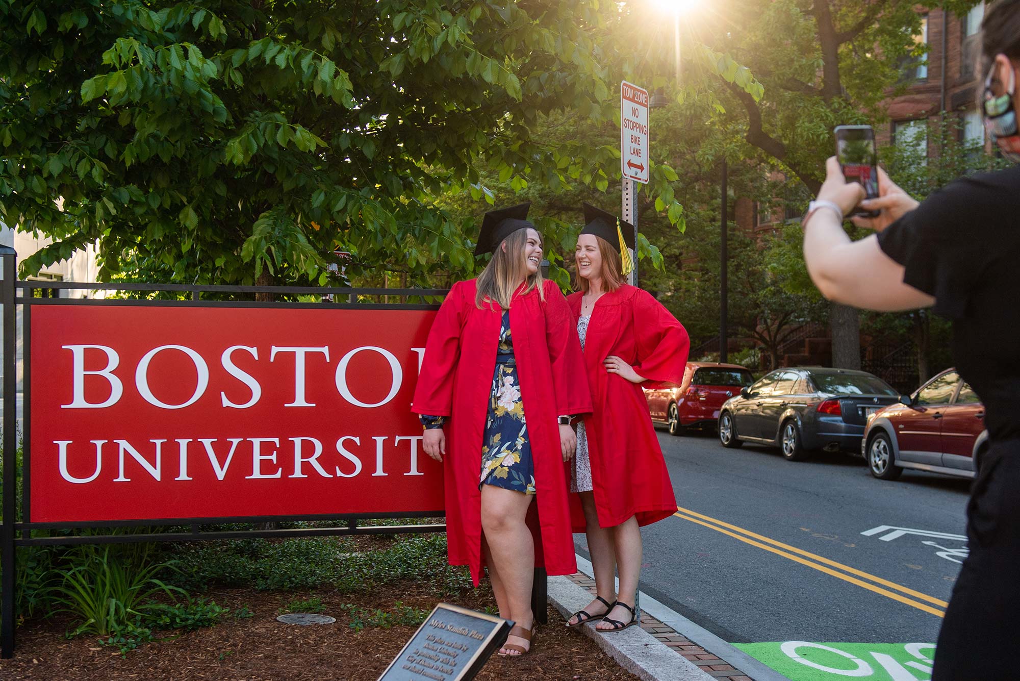 Photo of Claire Johnson (SAR’18, SSW’20, MPH’21), from left, and Kathryn Sparks (SSW’20) posing in their grad robes for Alex Ben-Jakob (SAR’17) (whose arm holding up a phone is seen in the right foreground) at the Myles Standish Plaza on May 26, 2020. Johnson and Sparks smile next to a big red "Boston University" sign.