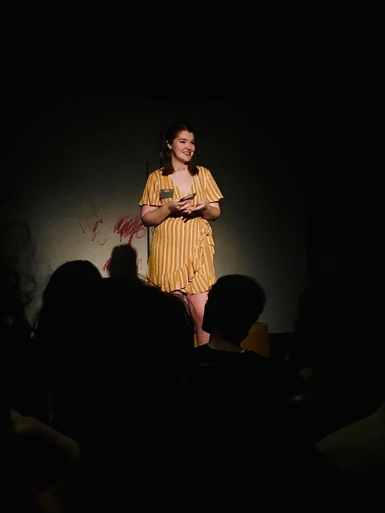 Photo of Abbey McCracken (Sargent’20) on stage in a mustard and white stripped dress. A spot light is on her and the silhouettes of heads are seen in the foreground.