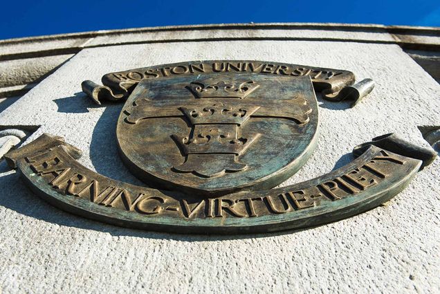 Photo of rusting, greenish gold BU seal on a stucco wall from underneath. Seal reads "Boston University, Learning, Virtue, Piety"