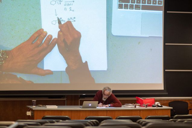 Photo of Bruno Rubio (GRS’96), a CAS master lecturer in chemistry, holding remote office hours for students in March. Rubio sits at a desk at the front of a lecture room and his hands working on a chemistry problem are seen on a projector screen behind him.