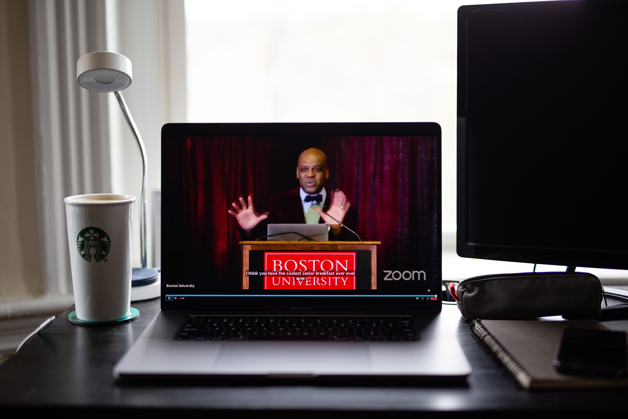 Photo of Sophie Park (CAS'20)'s laptop as she attends the Class of 2020's Senior Breakfast live stream from her bedroom on Fri. May 1, 2020. Laptop screen shows Dean Elmore speaking at a podium, a Starbucks cup sits to the left on the laptop. Photo by Sophie Park (CAS'20)