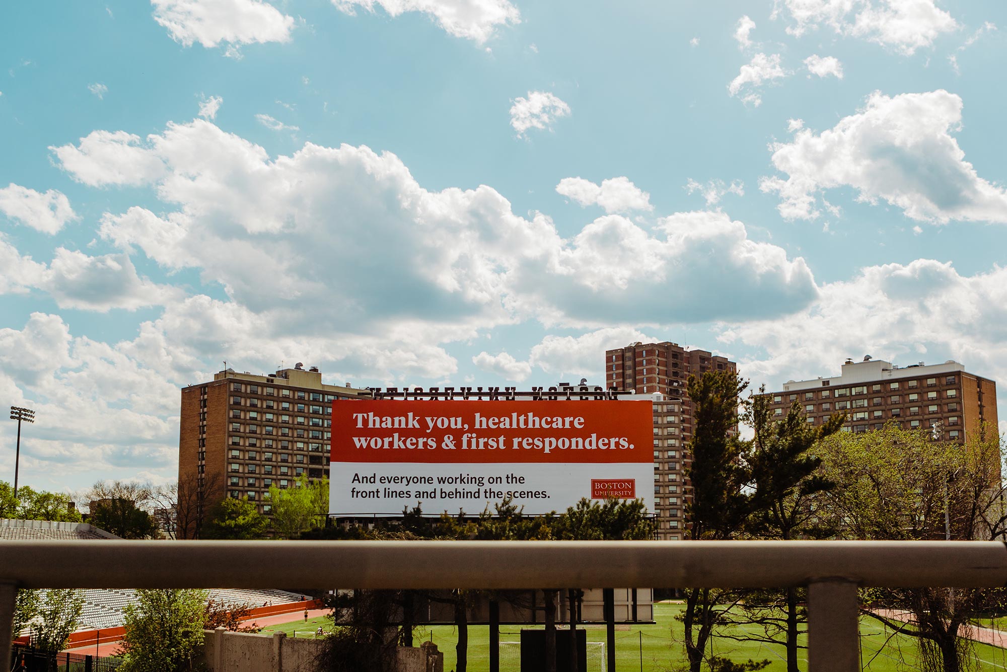 Image of a billboard behind Nickerson field thanks healthcare workers and first responders faces the Mass Pike. The sky is light blue with clouds. The sign reads "Thank you, healthcare workers & first responders. And everyone working on the front lines and behind the scenes.” The Boston University logo is seen on the bottom right of the sign.