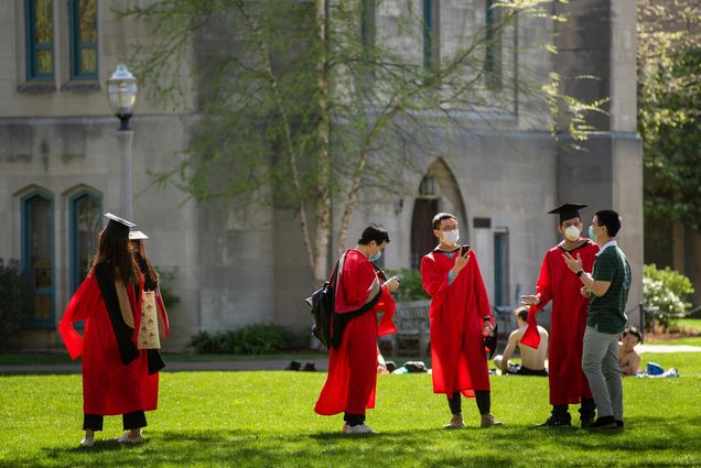 A group of students take graduation photos May 14, 2020. They stand in caps and gowns with masks on, and phones in hand, in the green space next to Marsh Chapel. 