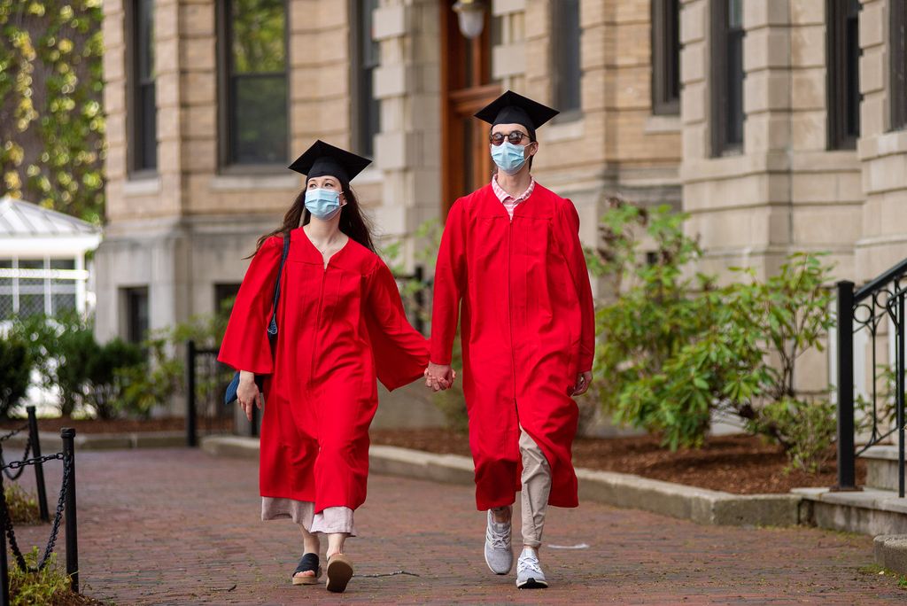 Joe Besser (QST’20) and Michelle Goltsman (COM’20) take a walk through campus May 16, 2020. Both wear blue face masks, red robes, and graduation caps, and hold hands as they walk.