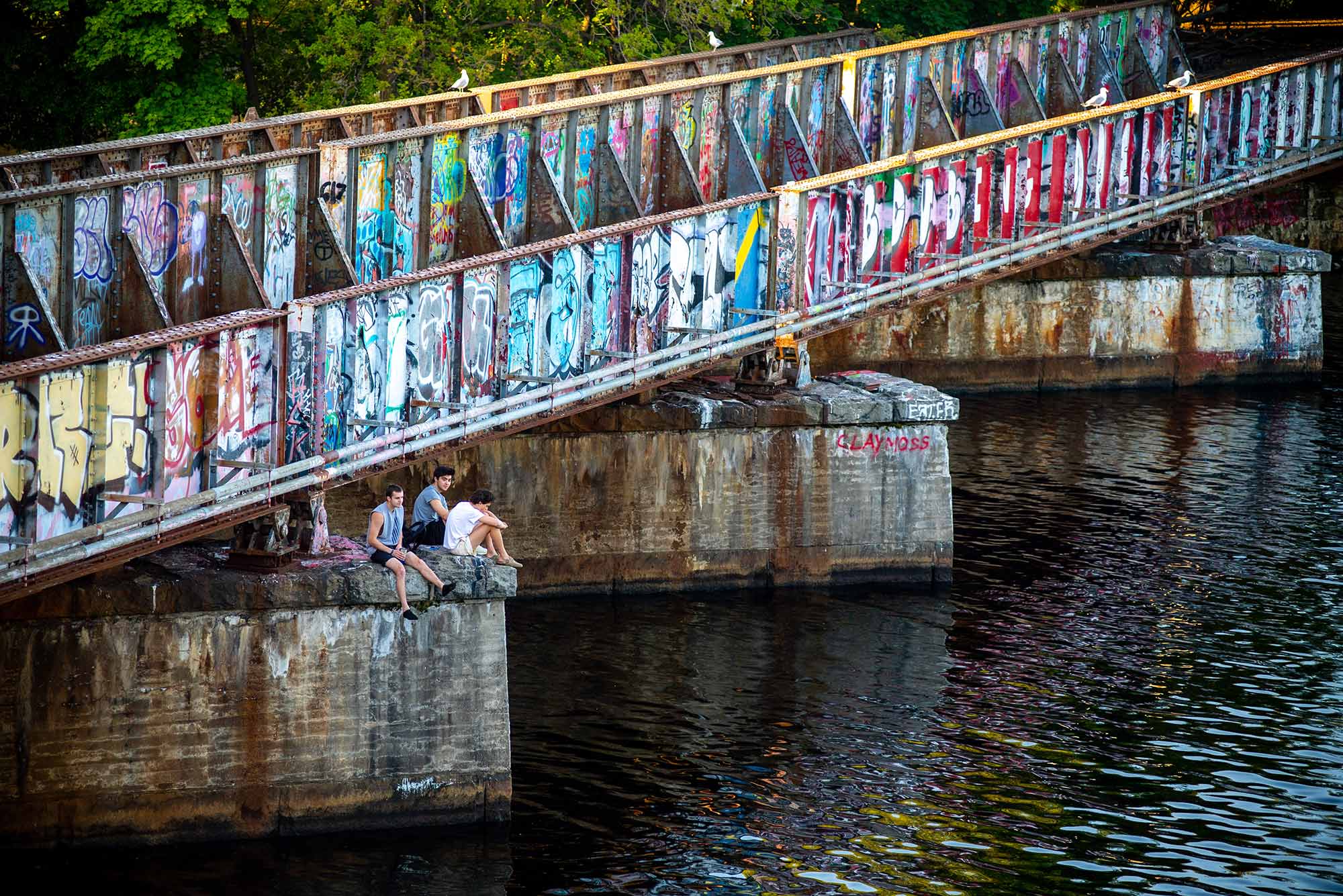 Photo of a small group of young people sitting underneath the BU bridge. the structure is covered with graffiti, the Charles River flows beneath it.