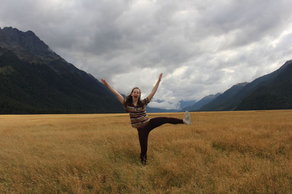 Photo of Michelle Goltsman (CGS’18, COM’20) with her hands and a leg in the air in a field with mountains in the background.