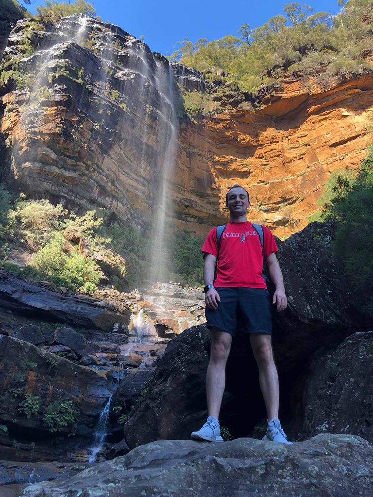 Photo of Joshua Vergules (Sargent’20) with a backpack on in front of waterfall.